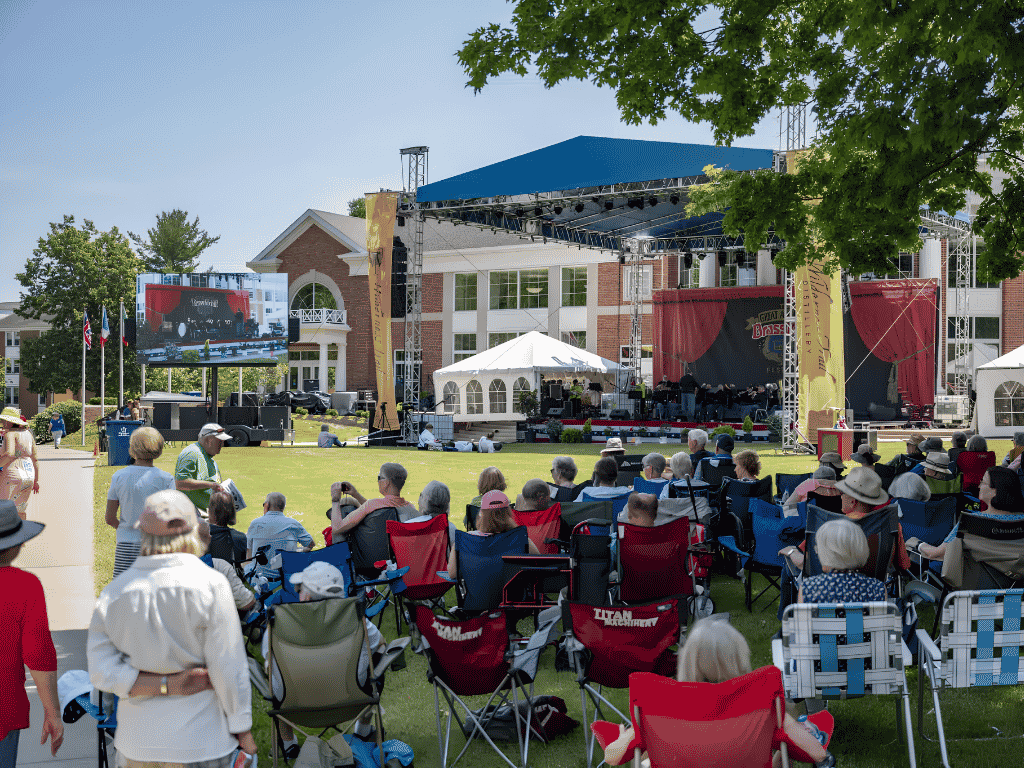 Outdoor community concert in a park with a large stage, blue canopy, and audience seated in colorful foldable chairs facing the performers.