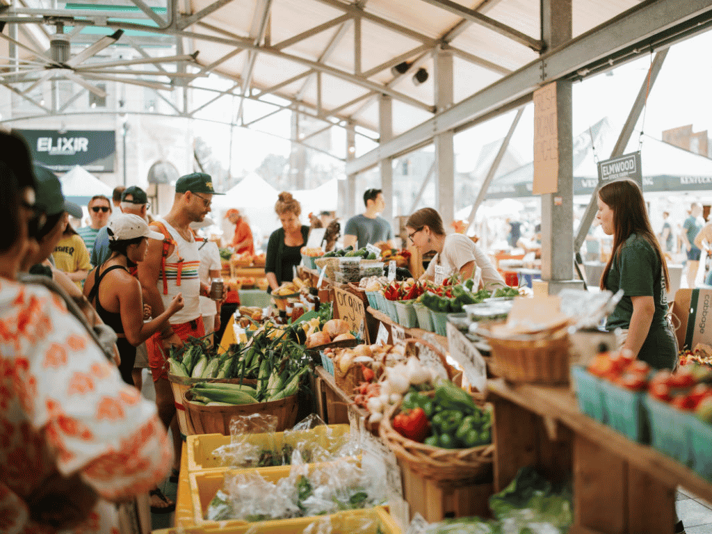 Busy indoor farmers market with shoppers selecting fresh produce from stalls under a metal roof.